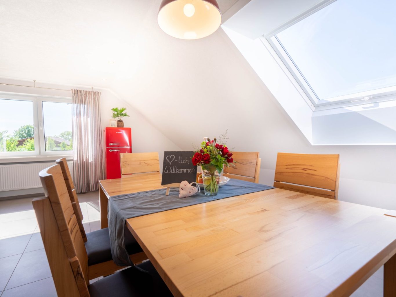Ferienwohnung im Schwarzwald Heller Essbereich mit Holz-Tisch, Stühlen und Blumenstrauß unter einem Dachfenster.